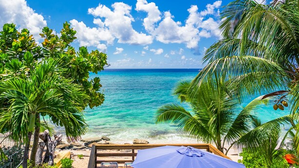 Vista de playa con una terraza de madera, sillas de descanso y una sombrilla azul.