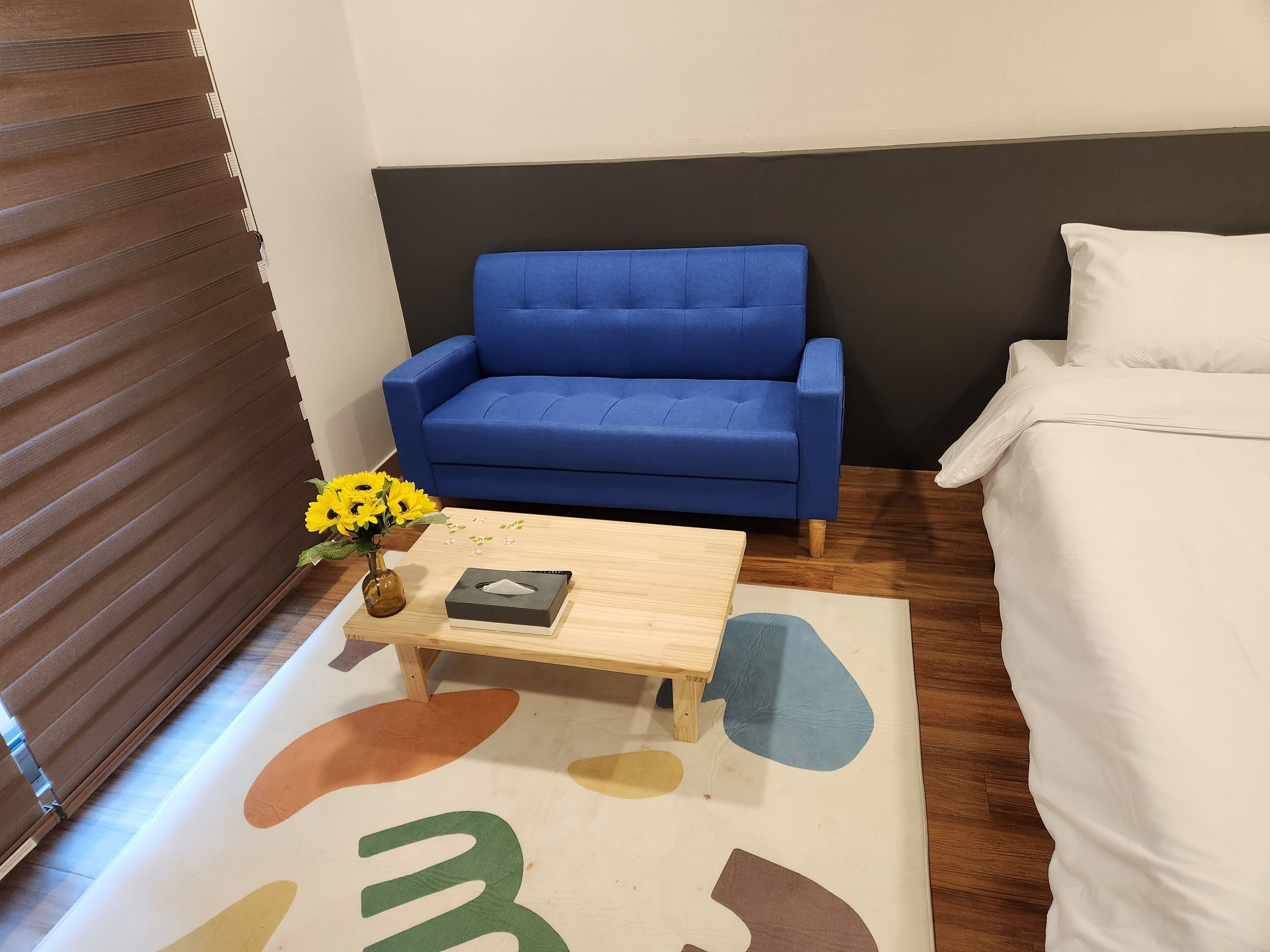 A small, modern room with a blue sofa, a white bed, and a wooden coffee table with a vase of yellow flowers.