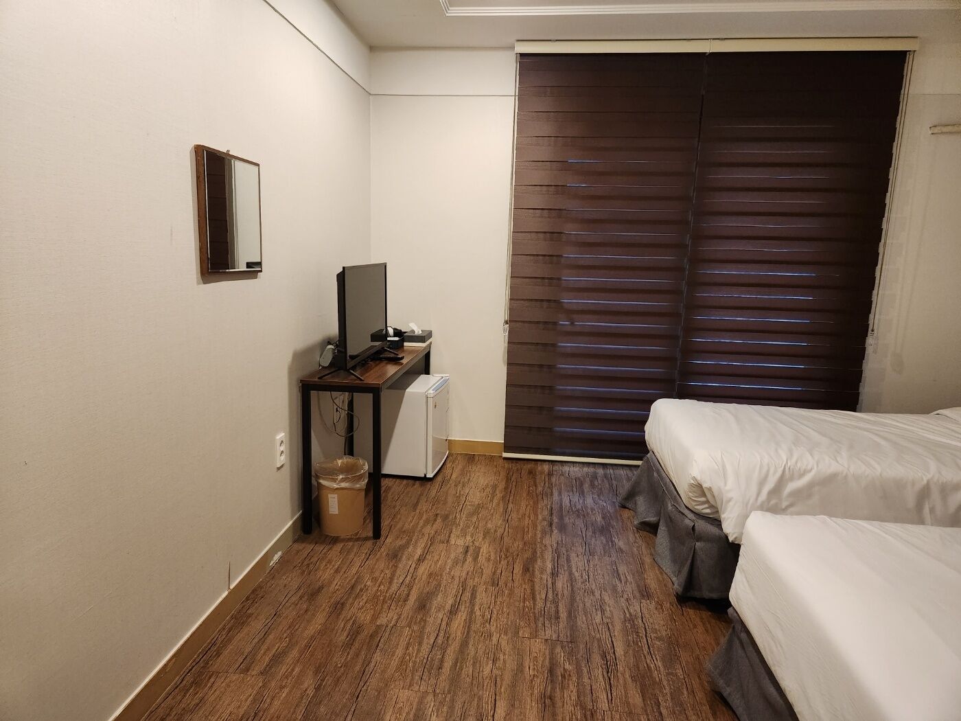 Habitació d'hotel petita amb terra de fusta, un llit, una taula de treball amb un ordinador, un petit radiador i una finestra amb persianes.