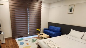 A hotel room with a bed, a blue sofa, a small table with a vase of flowers, and a window with blinds. - Hotel Navis (Jeju City)
