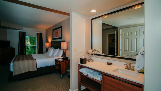 A hotel room with a bed, bedside table, lamp, and a large mirror. - The Suites at Hershey - 2 Bedroom (Hershey)