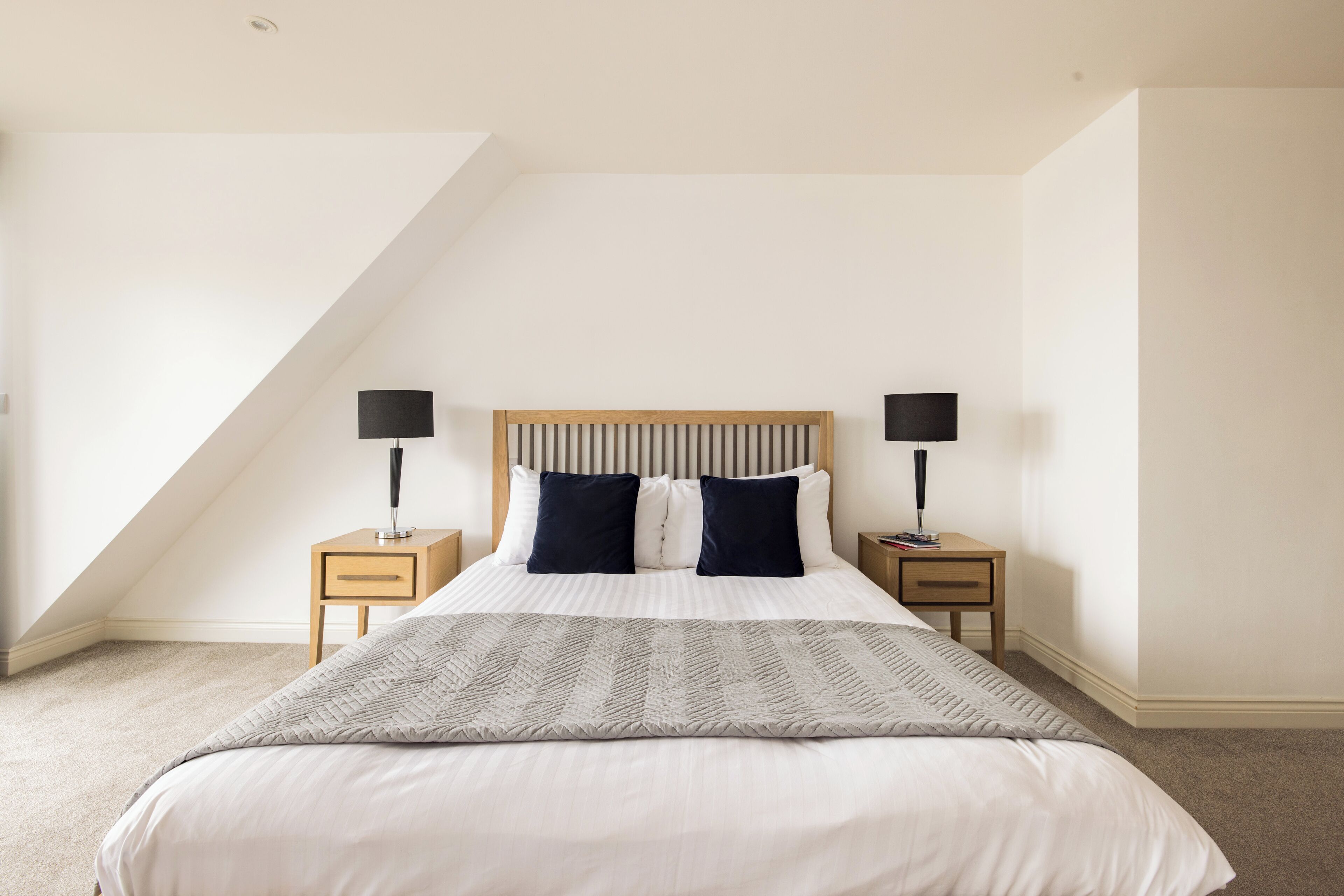 Quarto com teto inclinado, cama com cabeceira de madeira, duas mesas de cabeceira com abajures e uma janela grande.
