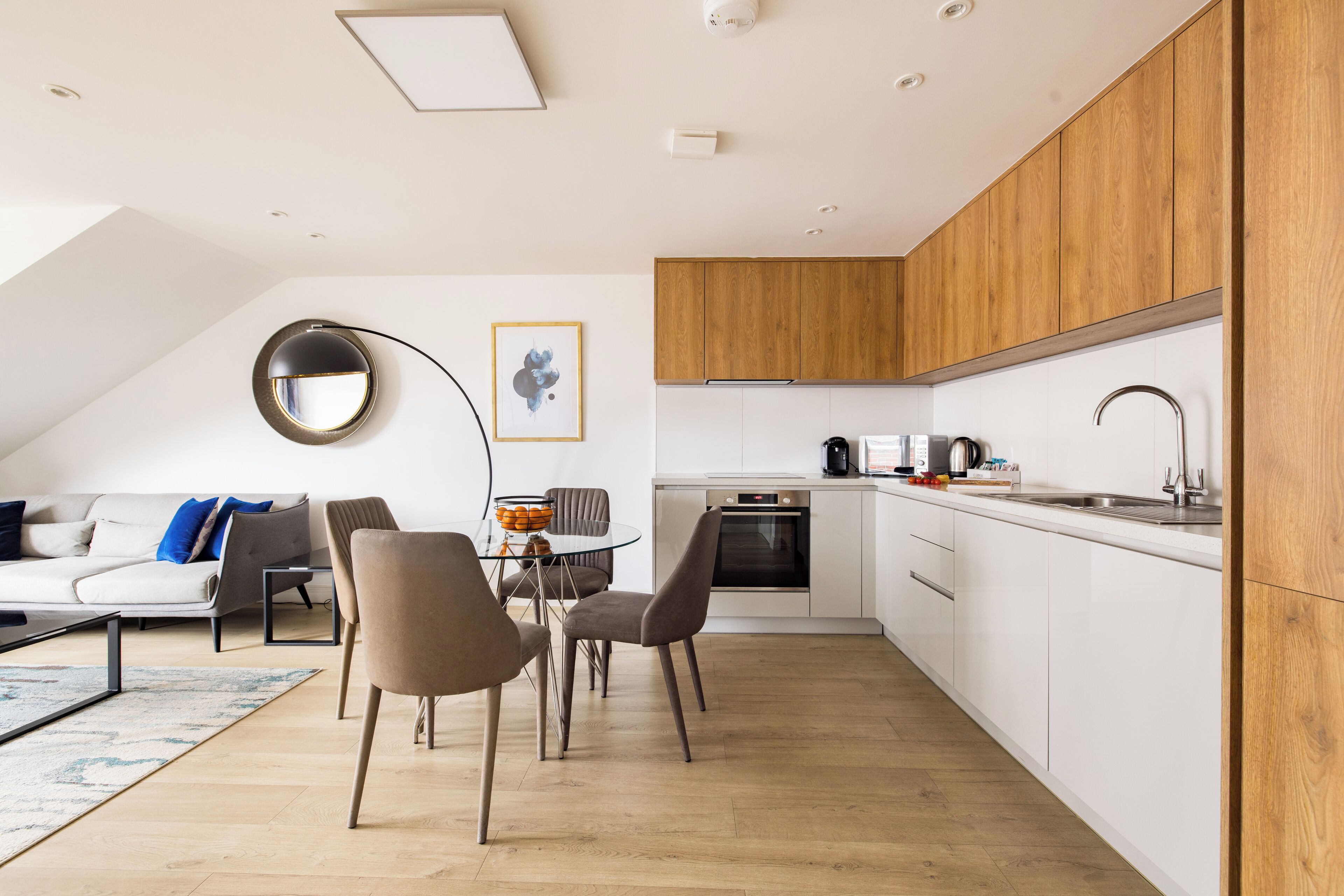 Eine moderne Küche mit weißen Schränken, einer Holzausziehtafel und einem eingebauten Ofen. Es gibt einen Essbereich mit Tisch und Stühlen sowie ein Wohnzimmer mit Sofa und Couchtisch.