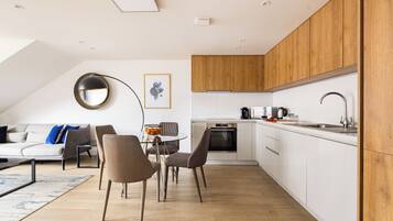 Una cocina moderna con armarios blancos, encimera de madera y horno empotrado. Hay una zona de comedor con una mesa y sillas, y una sala de estar con sofá y mesa de centro.