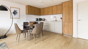 A modern kitchen with wooden cabinets, a dining area with chairs, and a black floor lamp. - Staines City Centre by Charles Hope (Staines)