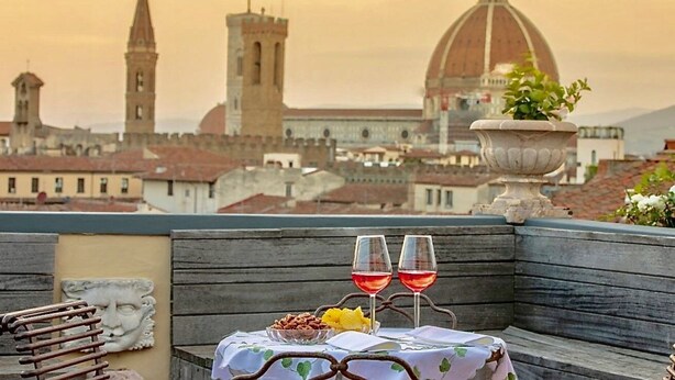 A table with a white tablecloth and two wine glasses on a rooftop terrace with a view of historic buildings and a dome in the background.