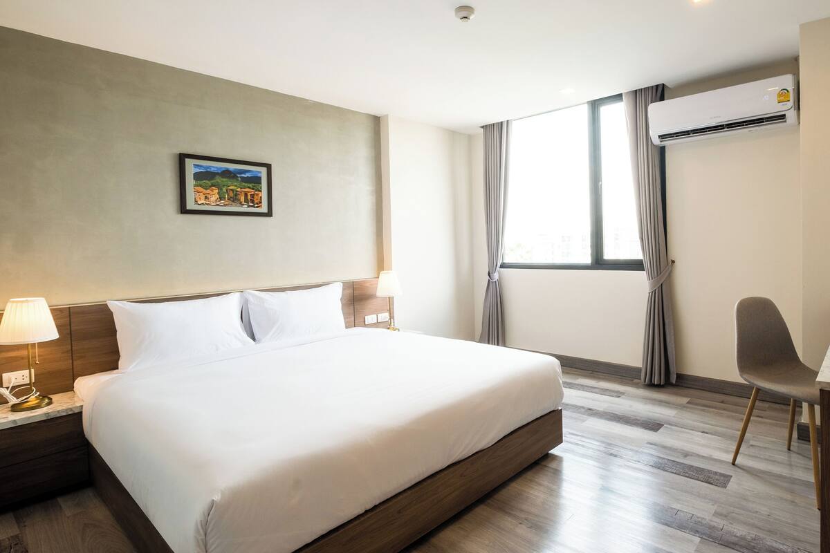 A hotel room with a large bed, a desk, a chair, a TV, and an air conditioning unit.