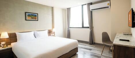 A hotel room with a large bed, a desk, a chair, a TV, and an air conditioning unit.