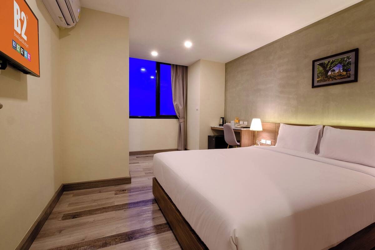 A hotel room with a large bed, a desk with a chair, a small table with a lamp, and a picture on the wall.