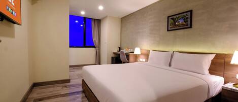 A hotel room with a large bed, a desk with a chair, a small table with a lamp, and a picture on the wall.
