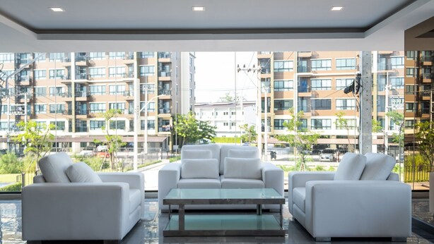 A modern living room with white sofas and a glass coffee table, large windows offering a view of a residential building complex. - B2 Korat Premier Hotel (Nakhon Ratchasima)