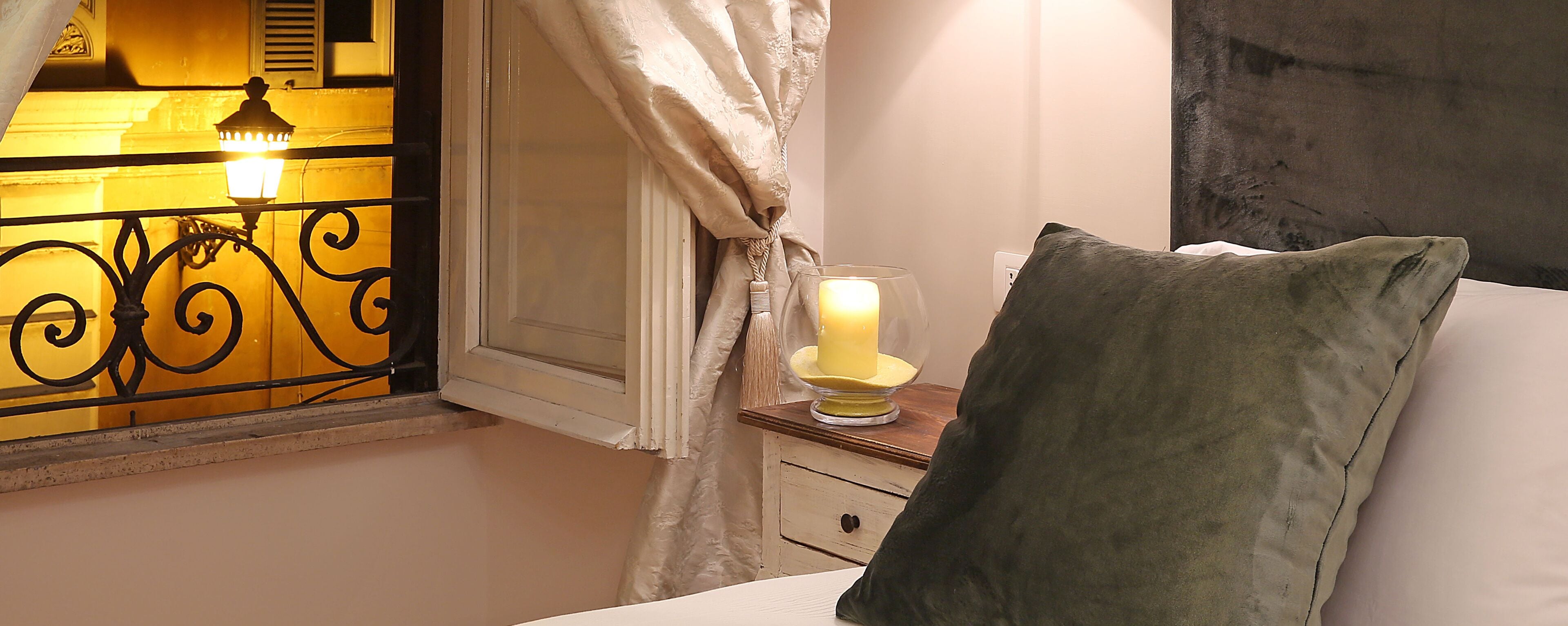 A bedroom with a bed, a bedside table with a candle, a window with curtains, and a wall-mounted light fixture.