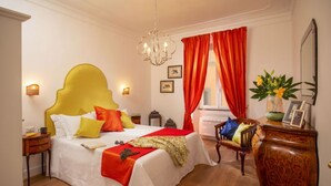 A hotel room with a large bed, a wooden nightstand, a chair, a dresser, a chandelier, and a window with red curtains. - Daplace - Flaminia Apartment (Rome)