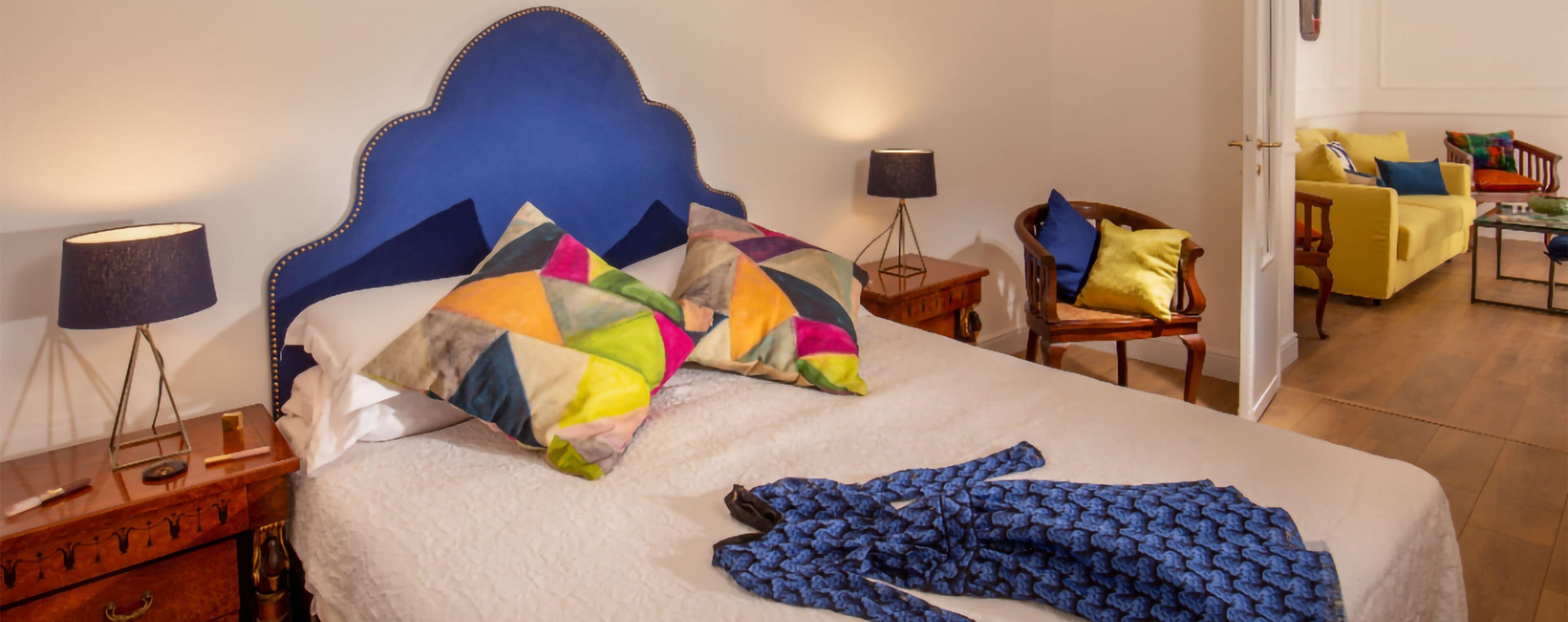 Een hotelkamer met een groot bed, een blauw hoofdeinde, een kleurrijke quilt, een blauwe deken, een houten nachtkastje, een lamp, een stoel en een gele bank.