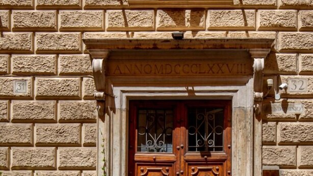 A building entrance with a stone facade, double wooden doors with metal handles, and a plaque above the door. - Daplace - Flaminia Apartment (Rome)
