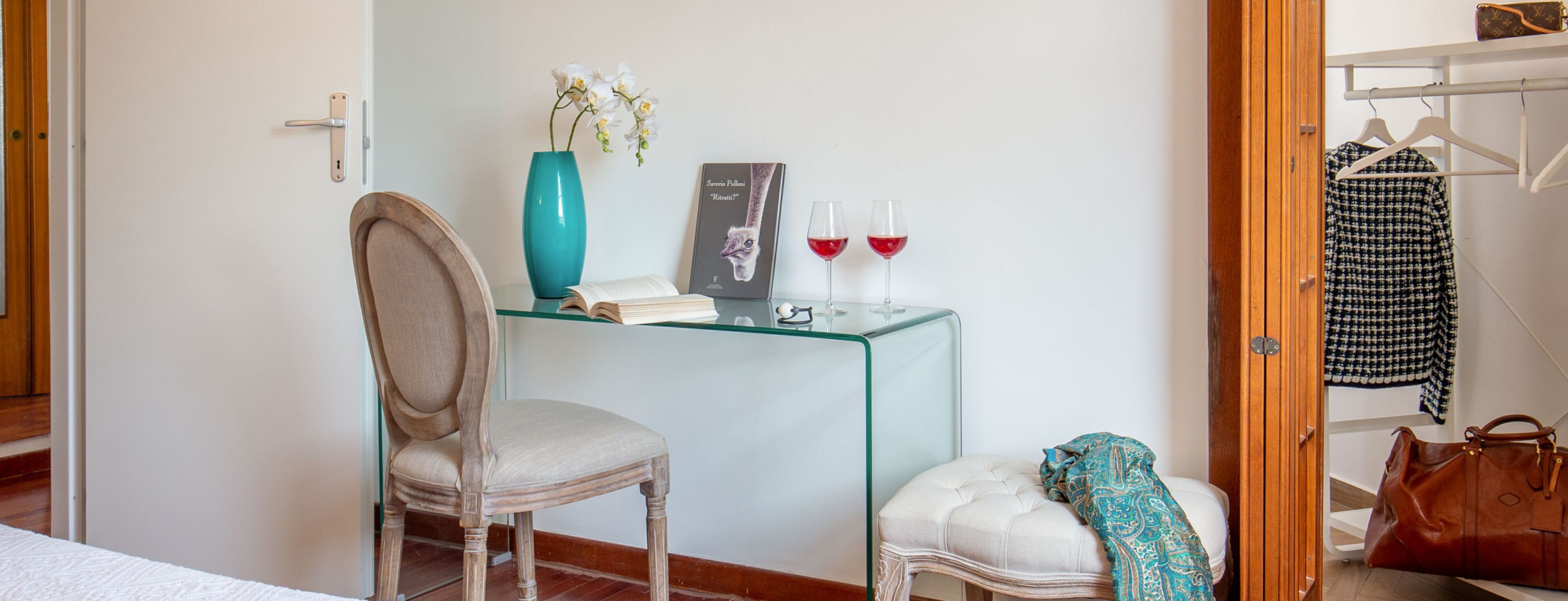 Ein Zimmer mit einem Holzstuhl, einem Glastisch mit Buch und Vase, einem weißen Ottomanen und einem Kleiderhaken mit einer Tasche.