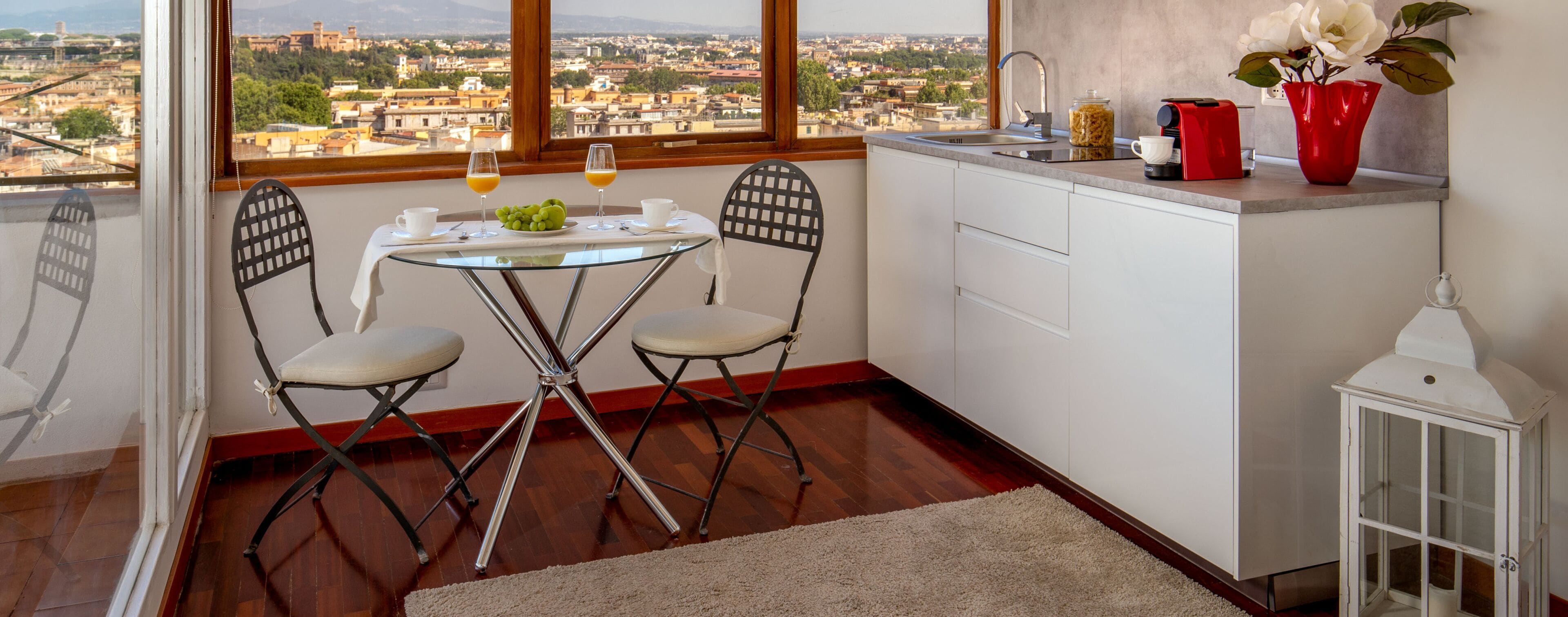 A small kitchen with a dining area, a view of the city, and a red vase with flowers.