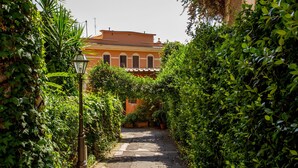A narrow alleyway flanked by lush greenery and a building with a red facade. - Daplace - Terenzia Apartment (Rome)