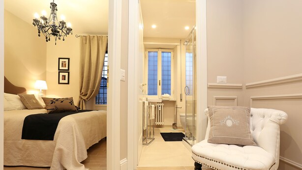 A bedroom with a bed, a chair, a chandelier, and a bathroom visible through an open door. - Daplace - Lucrezia Apartment (Rome)