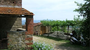 A rustic stone building with a tiled roof, a garden with flowering plants, and a parked scooter.