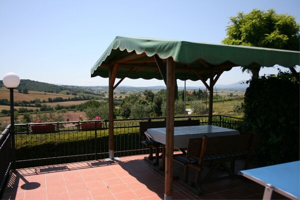 A terrace with a table and chairs, a pergola with a green awning, and a scenic view of hills and trees.