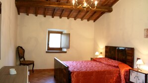 Un dormitori amb sostre de fusta, un llit amb estovatge vermell, una cadira, una taula petita, una finestra i una meseta de nit amb una làmpada.