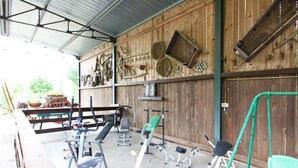 Ein überdachter Fitnessbereich im Freien mit verschiedenen Trainingsgeräten, darunter eine Hantelbank, Beinpresse und eine Klimmzugstange.