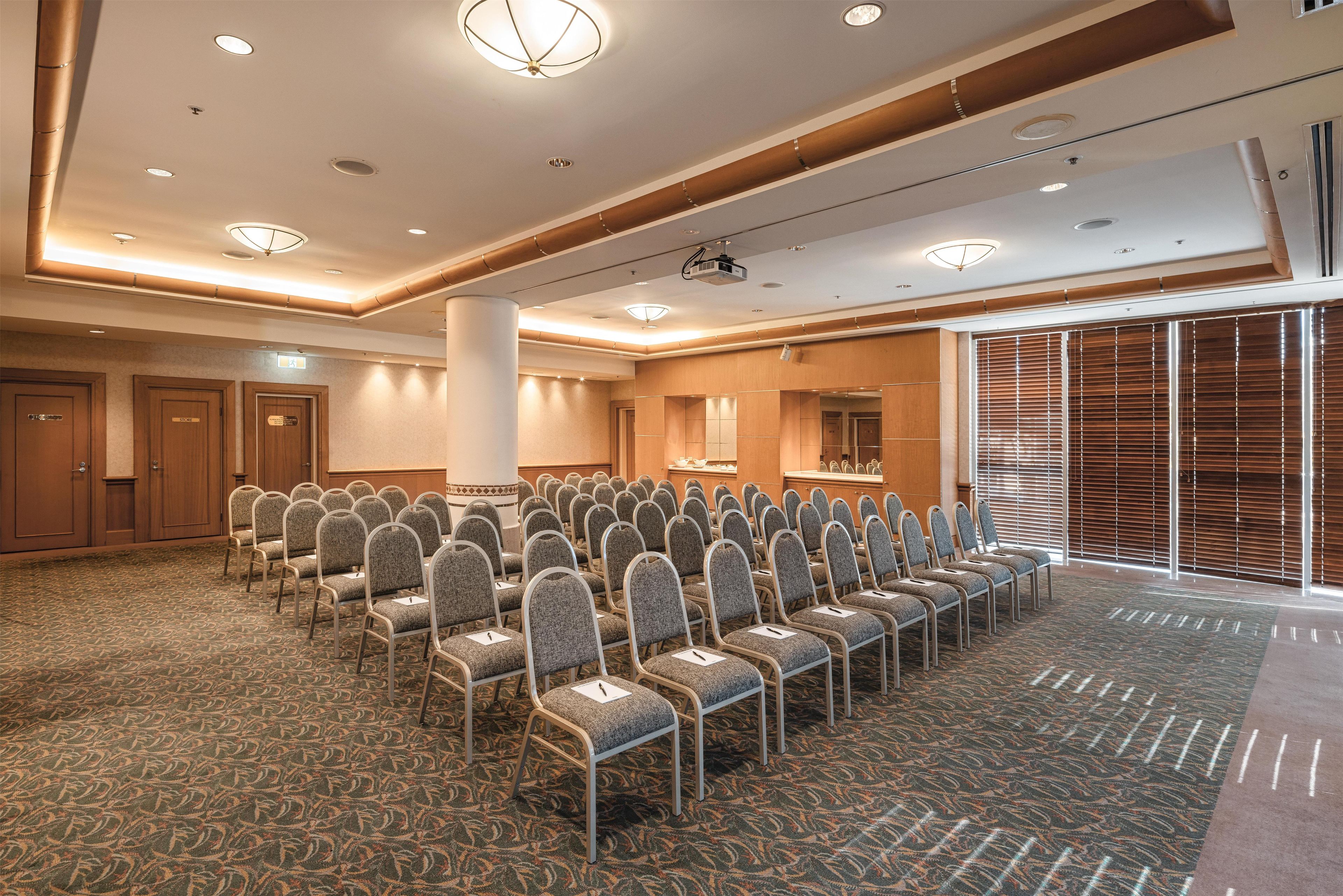 A conference room with rows of chairs, a stage with a projector, and a large window with blinds.