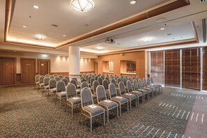 Een vergaderzaal met rijen stoelen, een podium met een beamer, en een groot raam met jaloezieën.