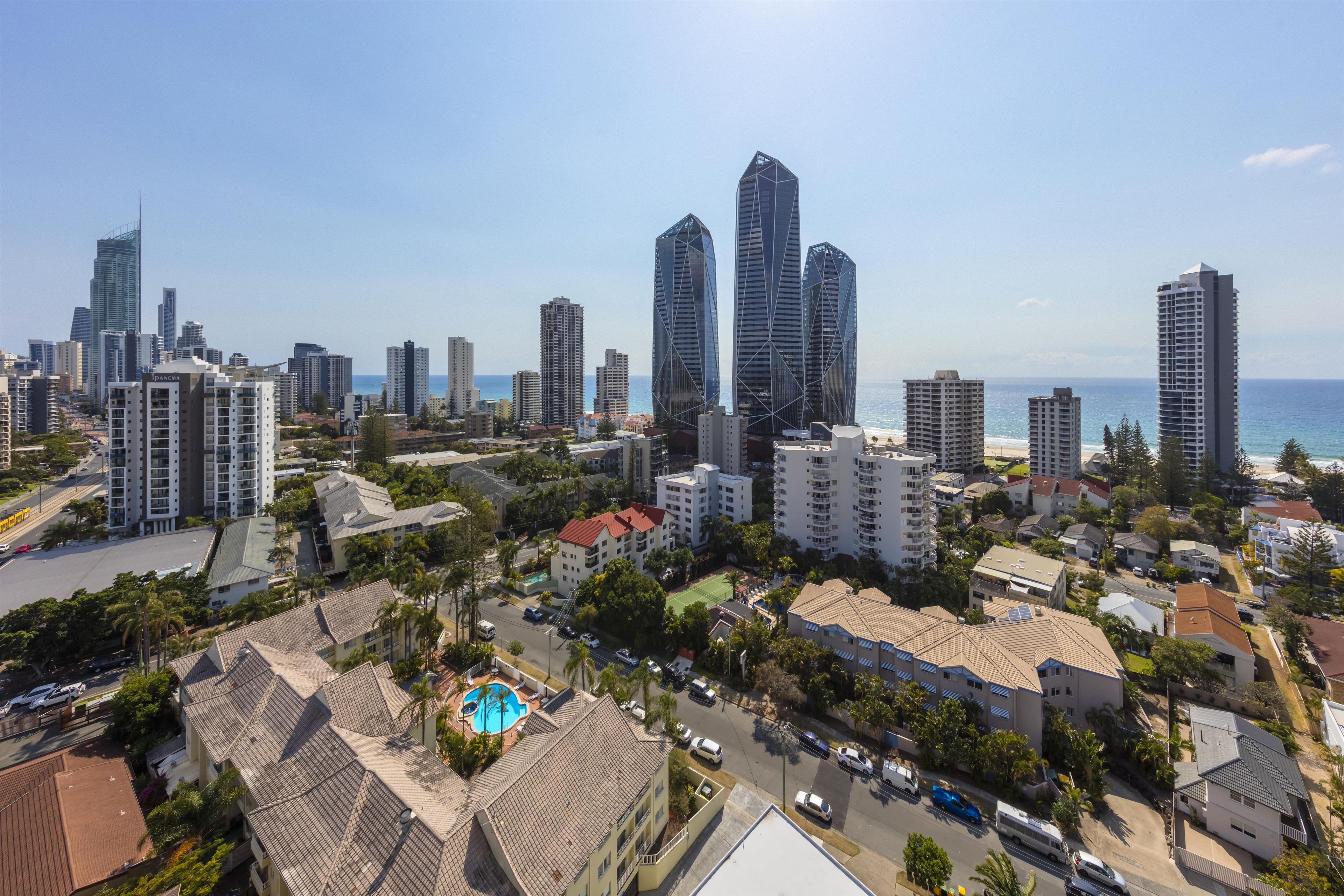 A coastal cityscape with modern high-rise buildings, a central swimming pool, and a clear view of the ocean.