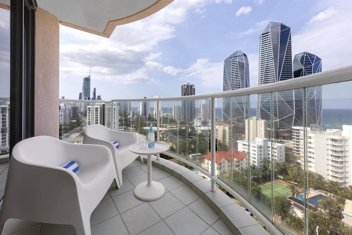 A balcony with white chairs and a small table, overlooking a cityscape with modern buildings and a clear sky.