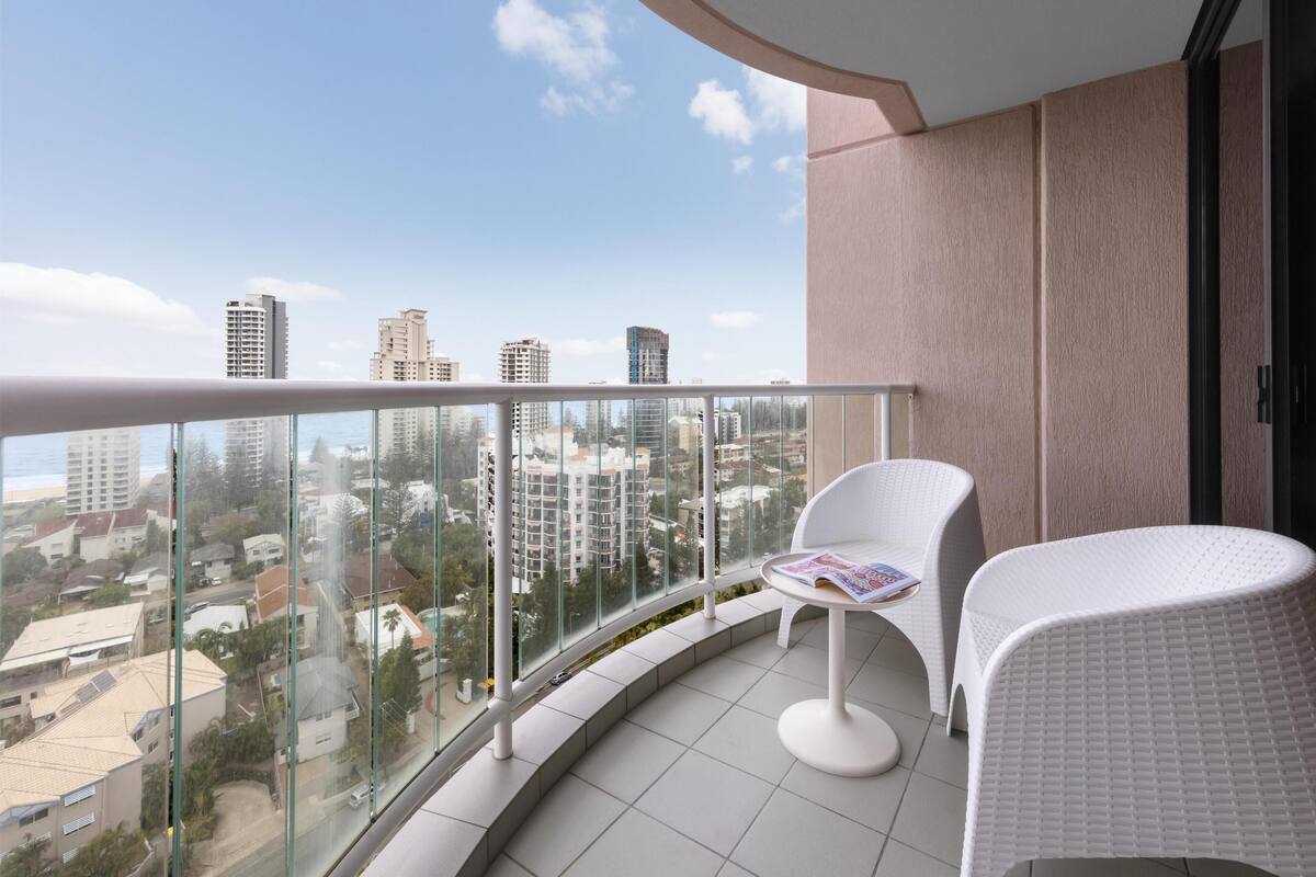 A balcony with a glass railing, white wicker furniture, and a view of a cityscape.