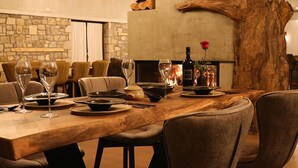 A dining area with a large wooden table set for a meal, surrounded by chairs. A fireplace with a log and a rose decoration is visible in the background. - Mylaon Boutique Hotel & Spa (Gortynia)