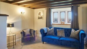 A room with a blue sofa, a chair, a lamp, a window with curtains, and a wall-mounted light fixture. - Host & Stay | The Coach House (Whitby)