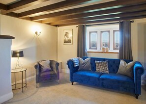 Una habitación con un sofá azul, una silla, una lámpara, una ventana con cortinas y una lámpara de pared.