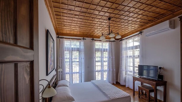 A bedroom with a wooden ceiling, a bed with white bedding, a desk with a television, and large windows with lace curtains. - Hotel Gorica (Berat)