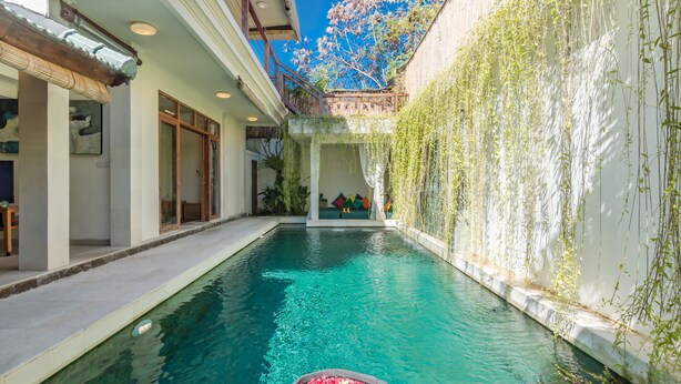 Una piscina con una balsa de piedra en primer plano, rodeada por un muro blanco con plantas trepadoras.