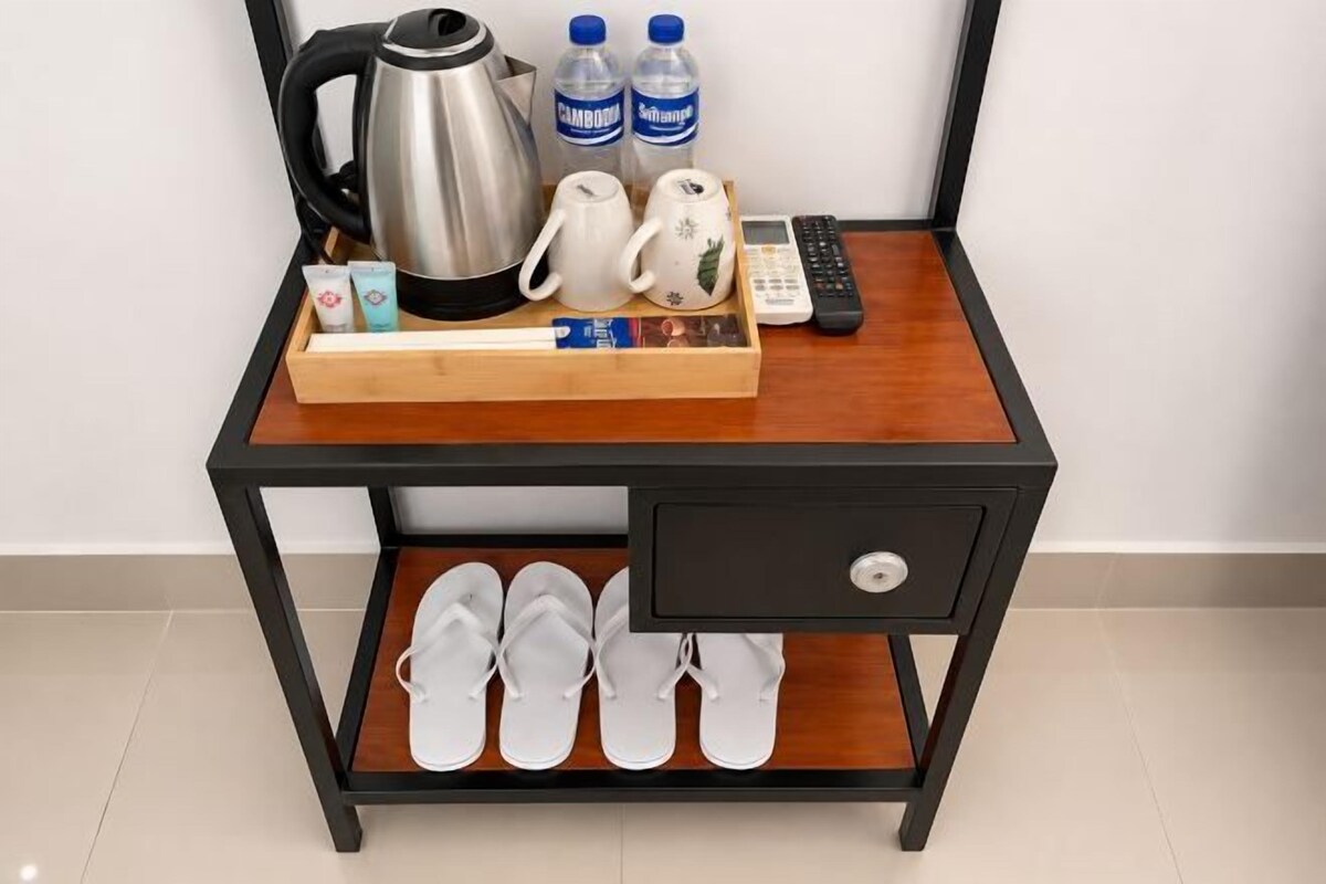 A wooden desk with a drawer, a tray with a kettle, bottles, a remote, and a cup.