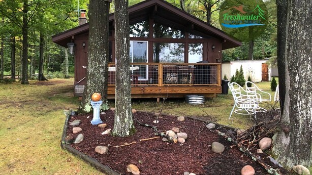 A cabin with a deck, surrounded by trees and a garden with rocks and a bird bath.