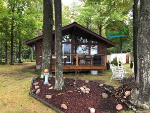 Une cabane dotée d’une terrasse, entourée d’arbres et agrémentée d’un jardin avec des rochers et un abreuvoir pour les oiseaux.