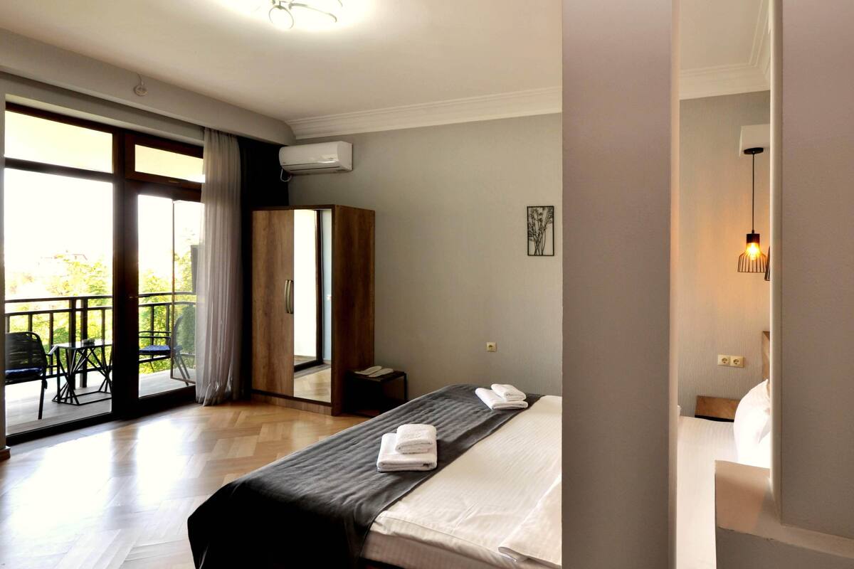 A hotel room with a bed, a wardrobe, a small table, and a balcony with a view of greenery.