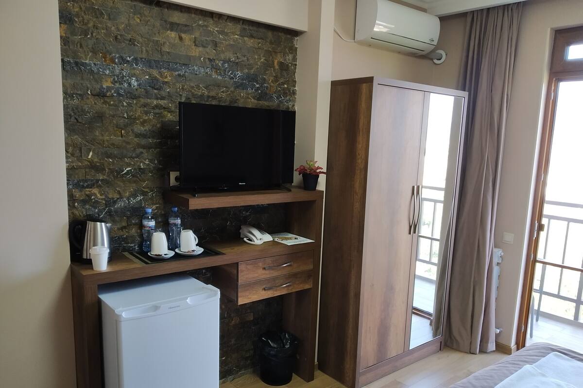 A compact hotel room with a stone wall, a flat-screen TV, a mini-fridge, a wooden wardrobe, and a sliding glass door to the outside.