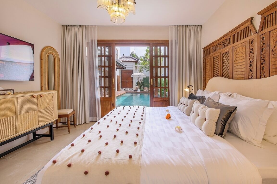 A spacious bedroom with a large bed, a wooden headboard, a bedside table, and a view of an outdoor pool area.