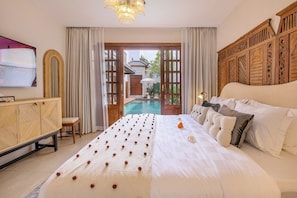 A spacious bedroom with a large bed, a wooden headboard, a bedside table, and a view of an outdoor pool area. - Villa Casa Adeline (Kerobokan)