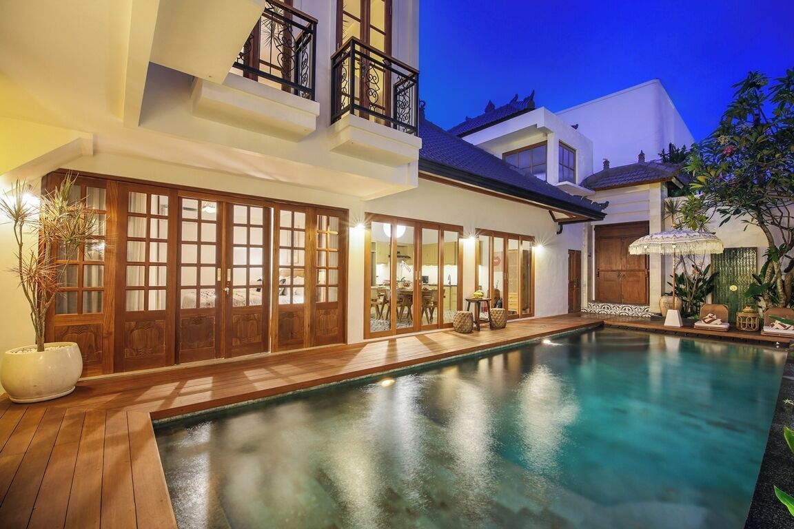 A two-story house with a swimming pool, wooden doors, and a balcony with black railings.