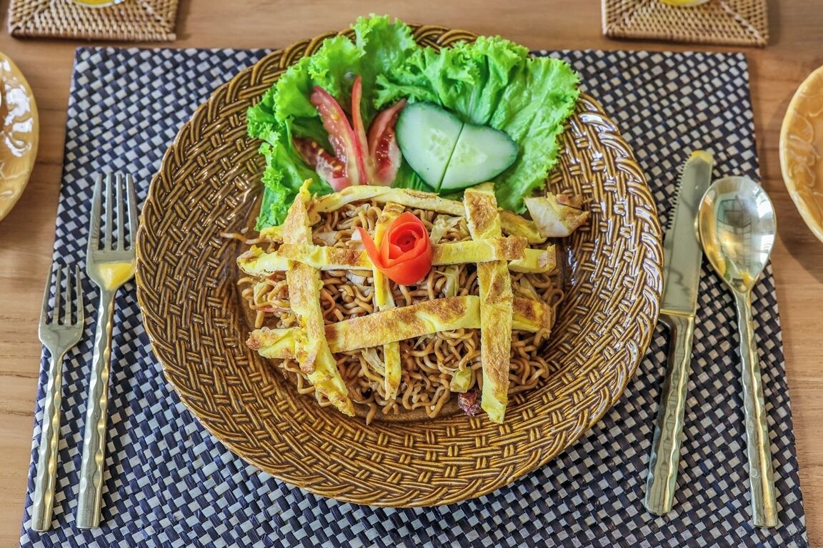A plate of noodles with vegetables and a decorative flower, served on a woven plate.