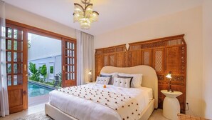 Un dormitorio amplio con una cama grande, cabecera de madera, mesita de noche con lámpara y vista a una piscina a través de una puerta abierta.