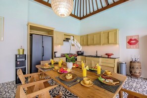 A dining area with a wooden table set for breakfast, a refrigerator, and a kitchen in the background. - Villa Casa Adeline (Kerobokan)
