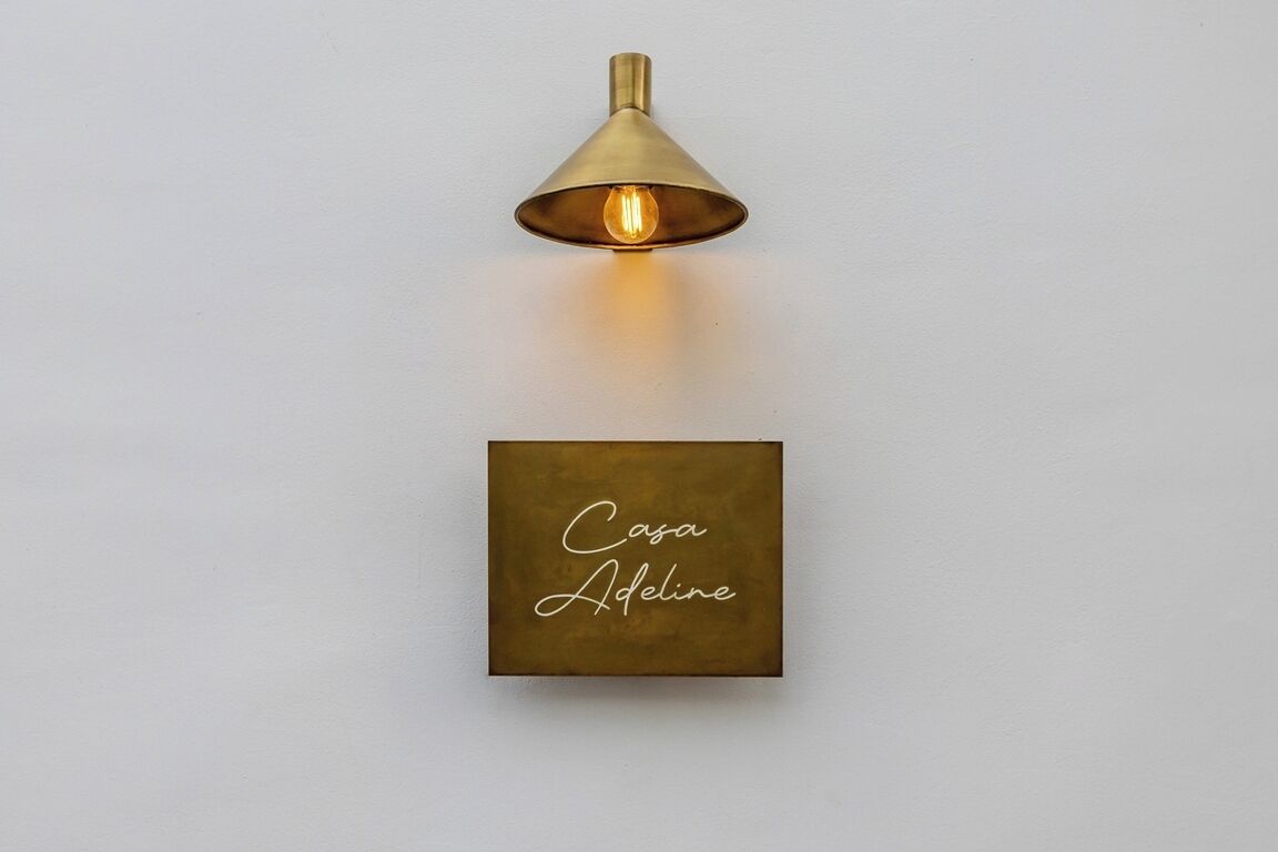 A wall-mounted light fixture with a conical shade and a lit bulb, and a square plaque with the text "Casa Adeline".