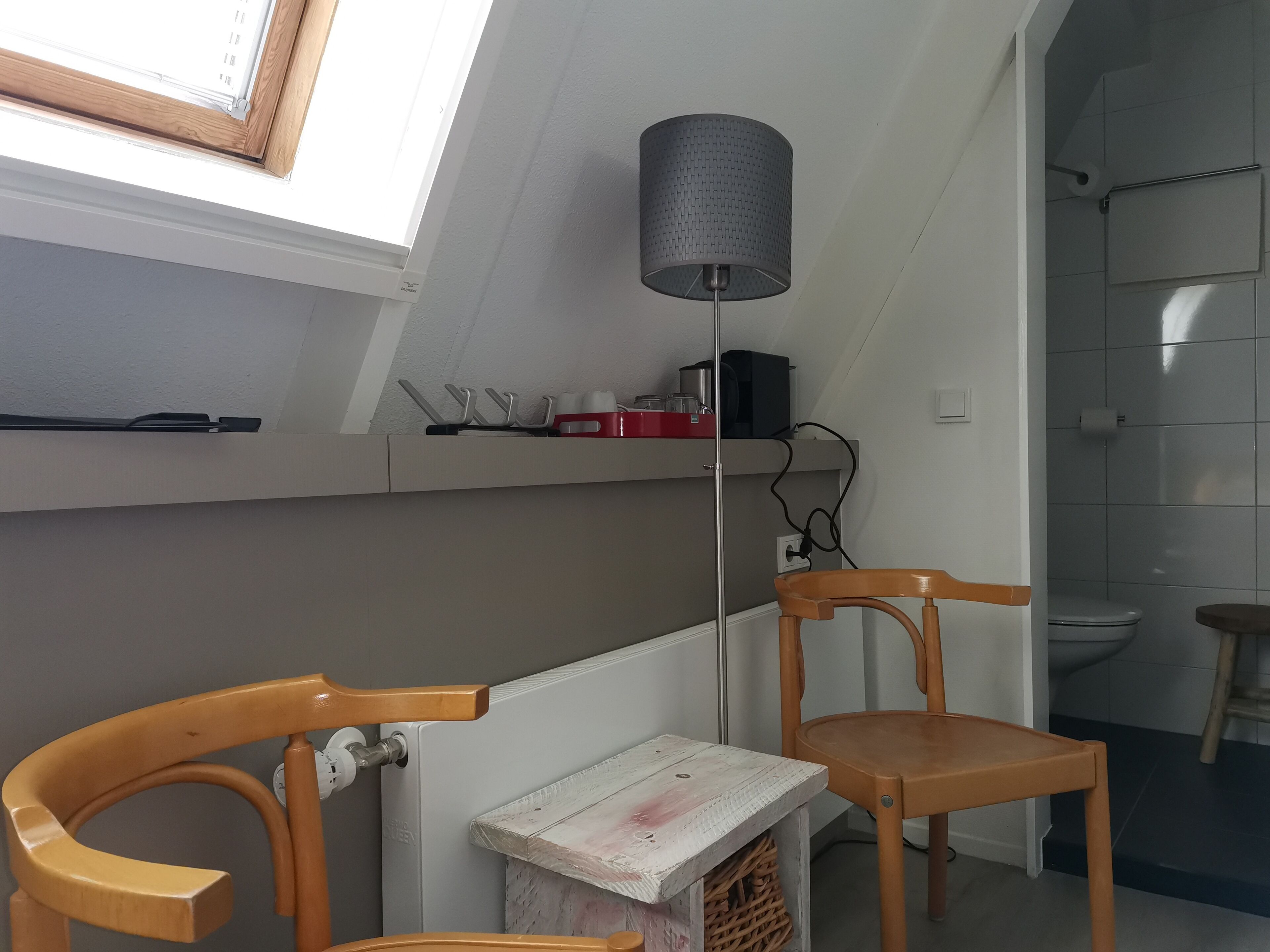 Une petite pièce aux dimensions réduites, avec un plafond en pente, un radiateur, une chaise en bois, une petite table et une étagère contenant divers objets.